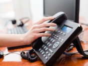 Hand greift zum Hörer eines Telefons auf einem Schreibtisch. Telefonhotline, Telefonberatung, telefonische Auskunft