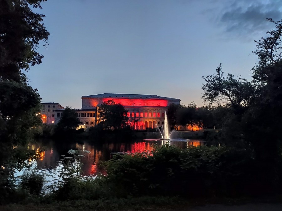 Zeichen zur Rettung der Veranstaltungsbranche: Night of light 2020. In Mülheim und Bundeweit wurden Gebäude mit rotem Licht illuminiert, um auf die dramatische Situation in der Veranstaltungswirtschaft, ausgelöst durch die Corona-Krise, aufmerksam zu machen. Das Foto zeigt den Blick auf die rot beleuchtete Stadthalle von der Ruhrpromenade aus. Davor die ebenfalls erleuchtete Wasserfontäne am Stadthallengarten auf der Ruhr. Im Vordergrund grüne Büsche. - Anna Moczurad/MST